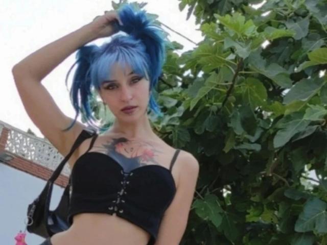 HannaDown, con cabello azul y esponjoso, sostén para el cabello, sosteniendo el cabello, mirando a la cámara, con un tatuaje en el pecho, un sostén negro, al aire libre, con un árbol de hojas verdes..