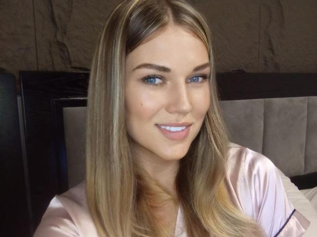 BernuLoverx has blue eyes, straight blonde hair, and a slight smile. She's wearing a pink satin shirt, showing her tits slightly. She's sitting on a bed.