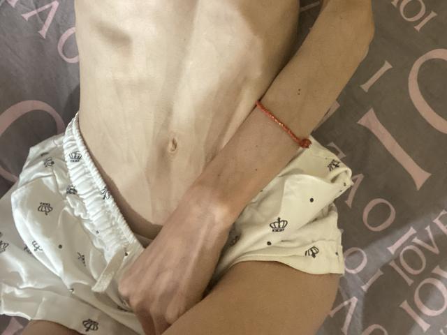 El torso delgado y pálido de Kabblasha, con las costillas visibles, lleva unos pantalones cortos blancos con motivos de corona, y tiene una pulsera roja en la muñeca derecha. Una mano cubre sus pequeñas nalgas desnudas..