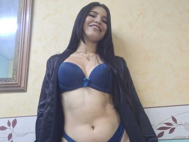 AmallaVega with long black hair, smiling, wearing a blue bra with a small bow and matching panties, black open robe, showing her tits and pussy, standing in a room.