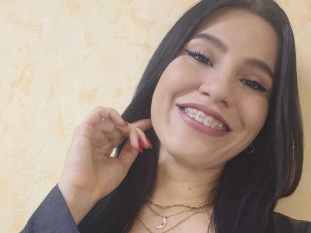 AmallaVega, with black hair and pink eyeshadow, smiles showing metal braces. She's touching her ear with red nails, wearing a black top and gold necklaces.
