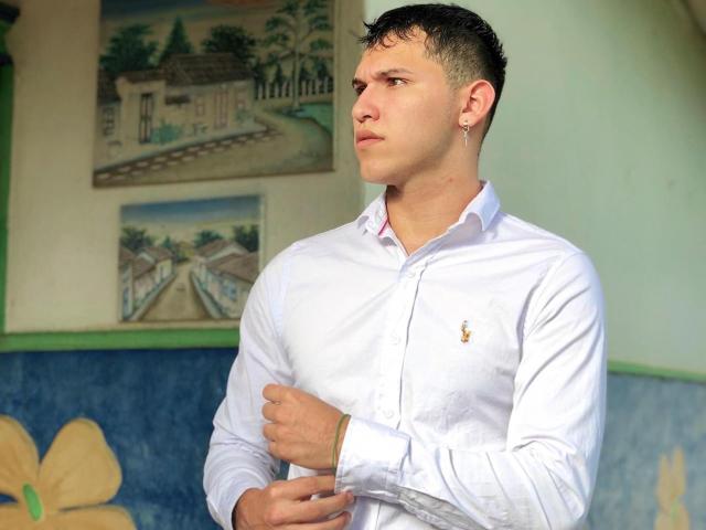 LorenzoReyes, white shirt, short black hair, small earring, looking to the side, right hand on chest, blue wall with yellow flower in background, slightly serious expression.