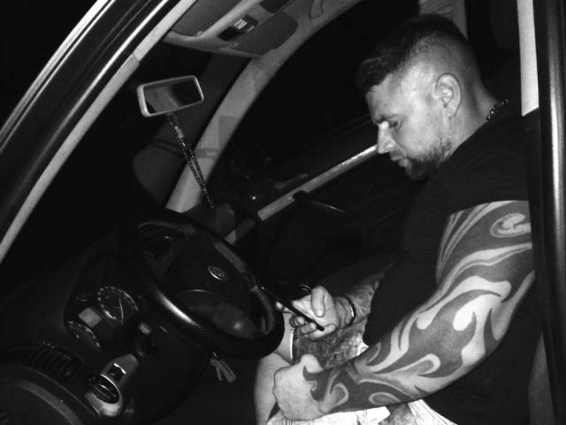 Black-and-white photo of KristianI with tattoos on his arm, sitting in a car, gripping a bag, looking down, dark short hair, beard, wearing a black shirt, night outside.