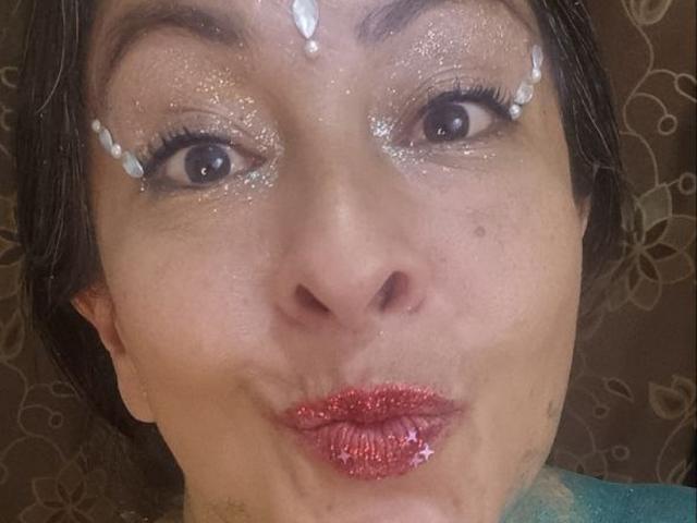 CatalinaJH's face with glittery eyes, white liquid dots on forehead and eyelids, and shiny red lips, puckering up. Brown hair, brown eyes, slight freckles, subtle makeup.