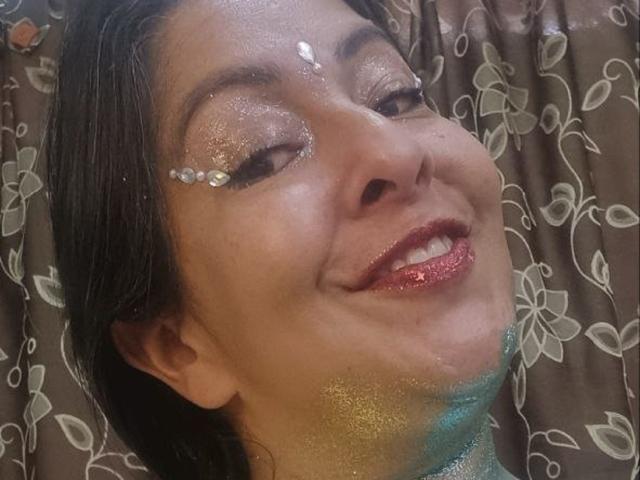 CatalinaJH with glitter on her eyes and mouth, wearing sparkly red lipstick, smiling up at the camera. She has dark hair and a brown curtain with white floral patterns.