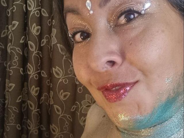 CatalinaJH's face is covered in glitter, with shiny gold and silver on her cheeks, forehead, and lips. She has red lipstick, and a leaf patterned curtain is in the background.