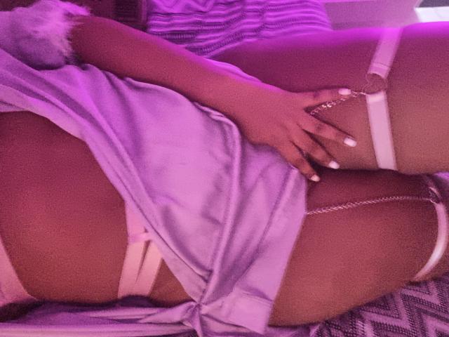 LunaMelynda-hot in a purple dress, hand on her ass, white straps around her thighs, pink-purple light, her hand has white nails, lying on a patterned surface.