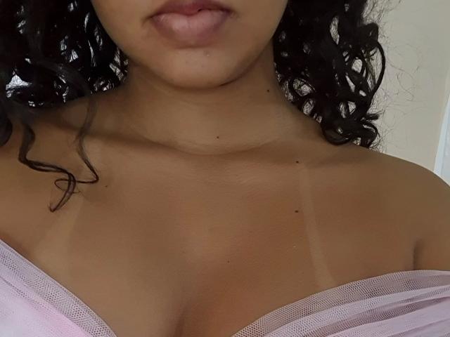 LunaMelynda-hot's tan, curly-haired face, pouty lips, and visible tan lines above her pink, sheer-clothed boobs. She has small moles on her chest and a smooth collarbone.