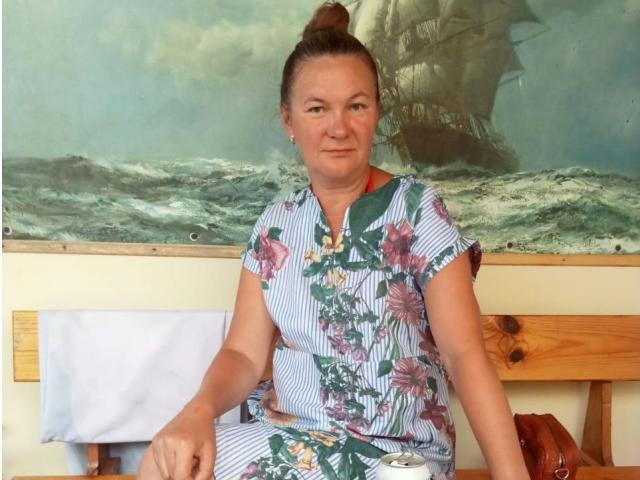 HolyMilfa with brown hair in a bun, wearing a floral blue and green dress, sitting on a wooden bench, white towel on left, brown bag on right.