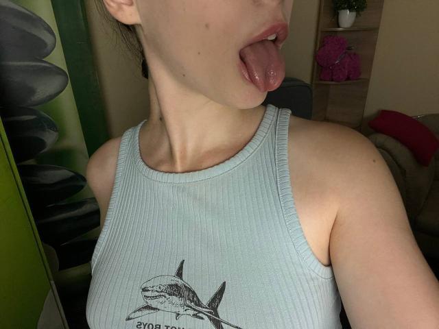 Valissia with her tongue out, mouth open, showing her pink tongue. She's wearing a light blue ribbed tank top with a shark graphic, and has small breasts.