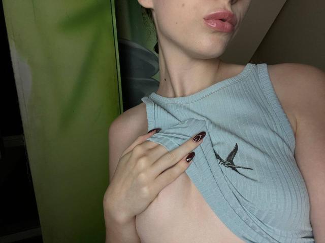 Valissia, met bleke huid, roze lippen en donkere nagellak, tilt haar lichtblauwe, geribbelde tanktop omhoog om haar kleine, onbedekte borst te laten zien. Op de top zit een klein plaatje van een vogel..