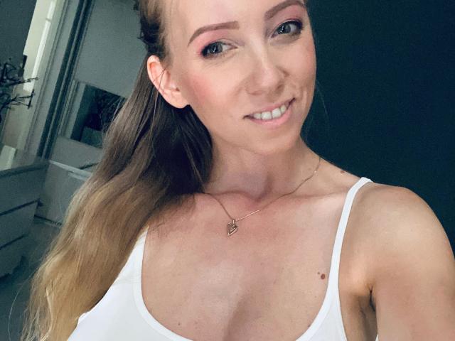 Julie Mayer – attraktiv mit langen braunen Haaren, rosa Lidschatten, einem weißen Tanktop, einer kleinen Herz-Kette, lächelnd, mit freiliegenden Brüsten, heller Haut, süß und einem kleinen Muttermal auf der Brust..