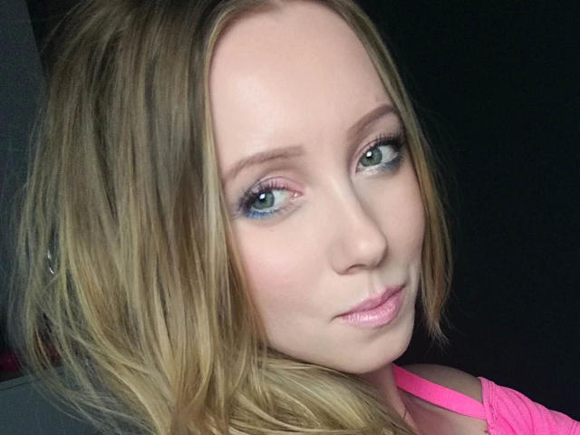 JulieMayer-hot a les cheveux blonds, les yeux verts et les lèvres roses, elle porte un haut rose, avec un maquillage discret et un léger sourire. Ses seins ne sont pas visibles sur cette photo..
