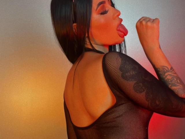 JunneLiu with long black hair, tongue out, wearing a black mesh top, showing her tattooed arm, and her back. Her tits are visible through the mesh.
