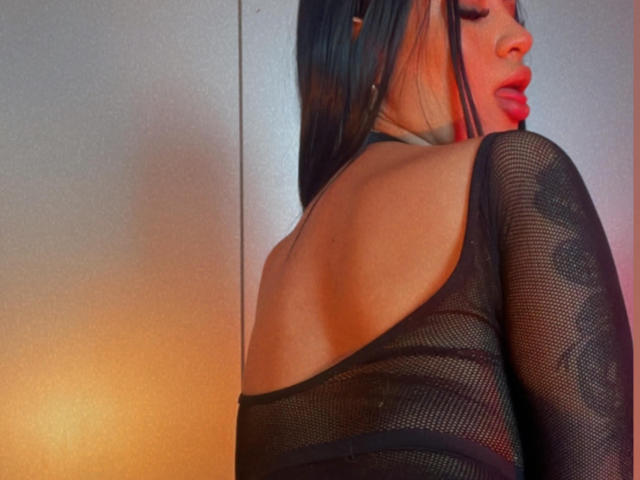 JunneLiu with long black hair, wearing a sheer black mesh top, exposing her back and upper buttocks, looking over her shoulder with red lips.