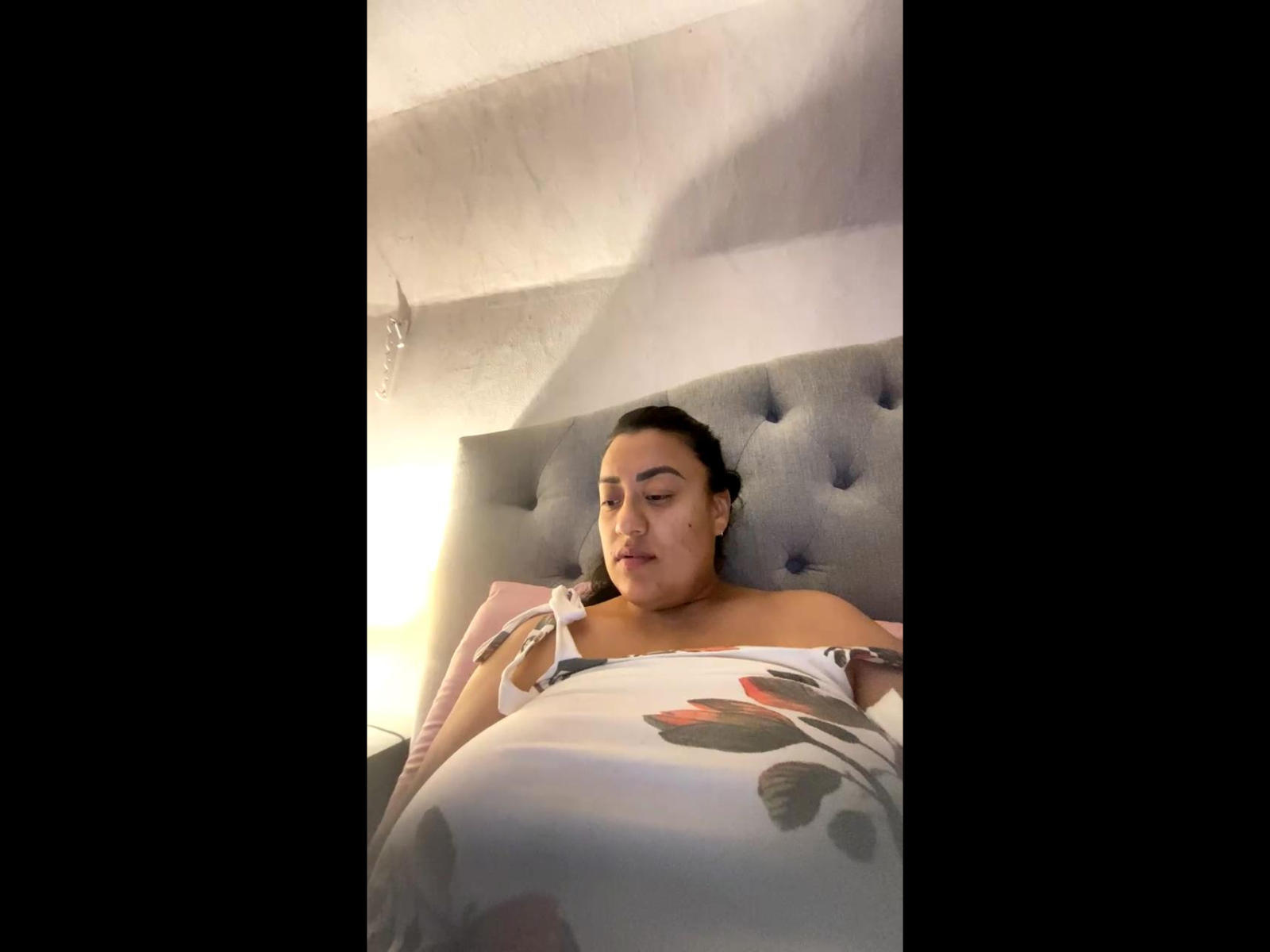 Alma69Hot is a pregnant woman with medium brown skin and dark hair, lying in bed with a floral-patterned blanket covering her tits and ass, eyes closed, in a dim room.