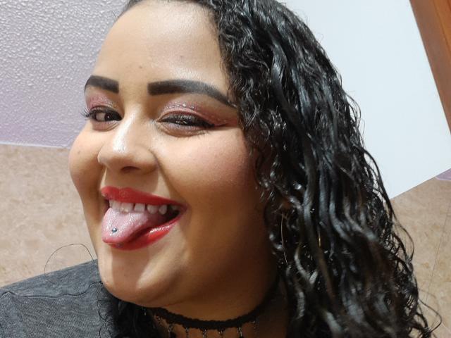 Melannie Carter ha i capelli neri e ricci, sopracciglia scure, ombretto rosso, rossetto rosso e un piercing sulla lingua. Sta sorridendo con la lingua fuori. Indossa una collana nera..