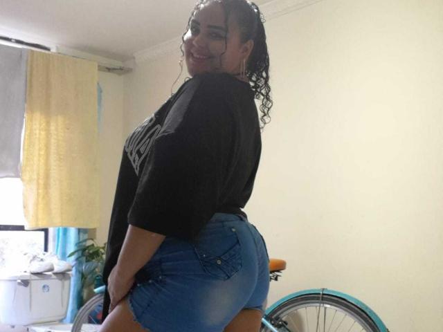 MelannieCarter with curly hair, smiling, wearing a black shirt and tight blue denim shorts, showing her big ass, standing in a room with a bicycle and yellow curtains.