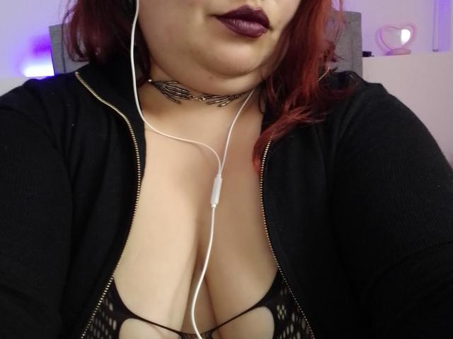 FoxyBell con capelli rossi, giacca nera con zip, scollatura profonda in rete nera, collana, auricolari bianchi, rossetto viola, leggermente sorridente, che mostra un seno generoso..