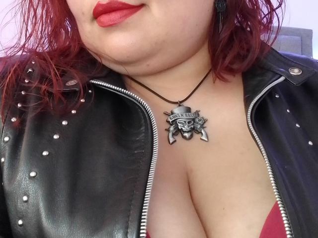 FoxyBell, avec du rouge à lèvres, une peau de couleur moyenne et des cheveux roux ondulés, porte une veste en cuir noir cloutée, qui révèle de larges seins, et un collier avec un crâne..