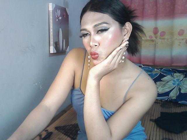 ErikaTS con labios fruncidos, vestido azul, mano en la mejilla, cabello oscuro en una coleta, maquillaje intenso, aretes, piel clara, sentada, dormitorio, piso de madera, almohada con estampado floral, cortina rosa..