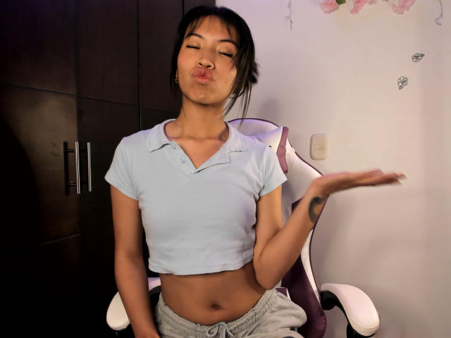 LuciaGarcia with puffy lips, wearing a light blue crop top and gray pants, sitting on a chair, hand raised, black hair, tattoo on right wrist.