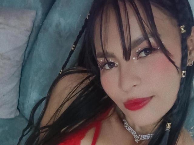 SusanKartier has black braids with gold beads, red lipstick, and a diamond choker. She's smiling, showing off her red lingerie, and has shiny eyes with sparkly makeup.