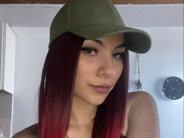 ValeryHarllow with red-dyed hair, green cap, and brown tank top, showing her nose ring and subtle smile, has tits visible above her tank top.