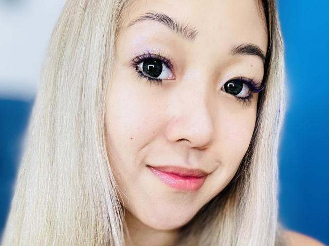 O rosto de RenyLime, com cabelo loiro, lábios rosa claro, olhos verdes e delineador rosa, sorrindo levemente, mostrando uma pequena pinta na bochecha direita..