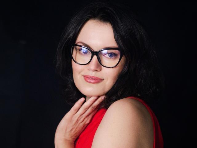 LolaHikls with black hair, wearing red sleeveless top, black glasses, and purple eye makeup, smiling softly, hand near chin, against dark background. Her tits are not visible.
