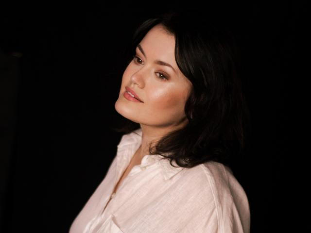 LolaHikls with shoulder-length black hair, wearing a white button-up shirt, has a subtle smile and slightly tilted head, looking softly at the camera against a dark background.