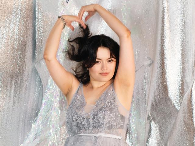LolaHikls with arms raised, wearing a sheer gray lace dress, exposing her tits, standing against shimmering silver fabric, black wavy hair, light skin, subtle smile, bracelet on right wrist.