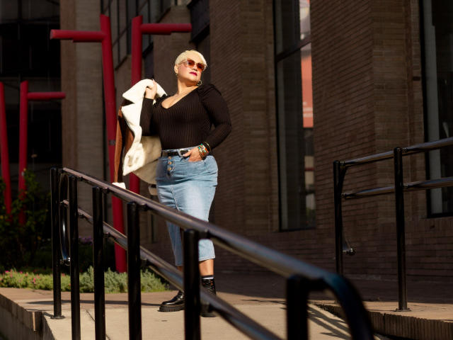 GianellaCox with blonde hair, wearing a black top, blue jeans, black shoes, and sunglasses, holding a white jacket, standing on a sunlit brick staircase.