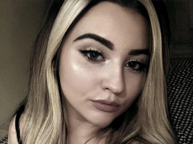 KamillaAss has blonde and brown hair, thick eyebrows, long eyelashes, and a neutral expression. Her face is slightly shiny with a small mole under her nose.