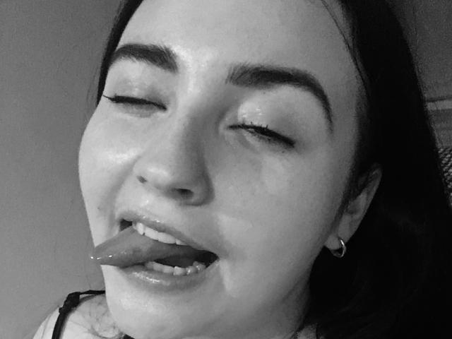 KamillaAss has her eyes closed with her tongue out, her wet face glistening. She has dark eyebrows, a hoop earring, and is wearing a black top.
