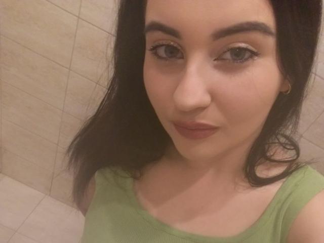 KamillaAss with dark brown hair, green eyes, and light skin wearing a green top, showing a subtle smile, and small hoop earrings, in a selfie.