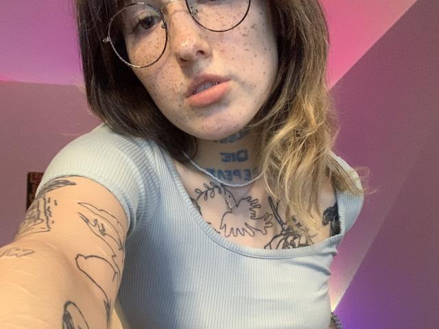 Ragazza con lentiggini, che indossa occhiali, maglietta a coste azzurra, con tatuaggi visibili sul petto e sul braccio, capelli biondi tendenti al castano, sfondo rosa, labbra leggermente dischiuse, selfie della parte superiore del corpo..
