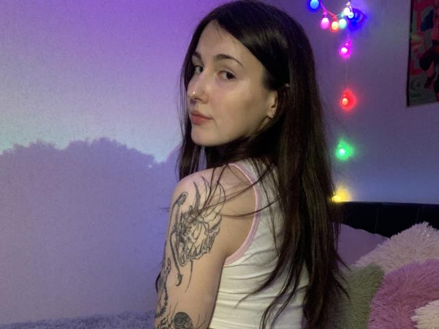 SweettSoft met lang bruin haar, een witte tanktop en tatoeages op haar rechterarm, kijkt over haar schouder, in een kamer met kleurrijke lichtslingers en een pluizig kussen..