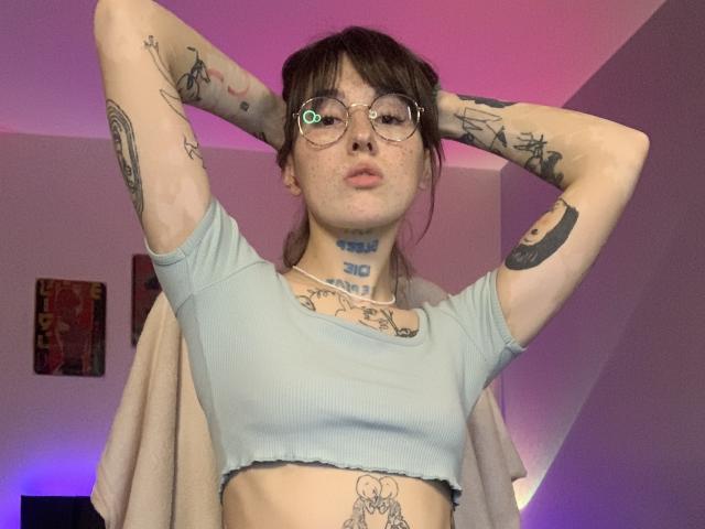 Una chica con tatuajes, SweettSoft, con gafas, labios fruncidos y pecas, vestida con una camiseta corta azul claro, con las manos detrás de la cabeza, mostrando los tatuajes y el piercing en el ombligo, en una habitación iluminada con luz rosa..