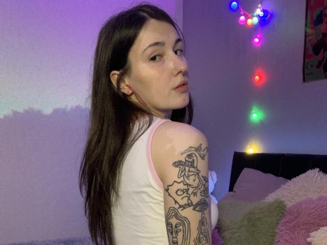 SweettSoft, avec ses longs cheveux bruns, son débardeur blanc et ses tatouages sur les bras, regardant par-dessus son épaule, une lumière violette sur le mur, des guirlandes lumineuses colorées et des coussins moelleux en arrière-plan..