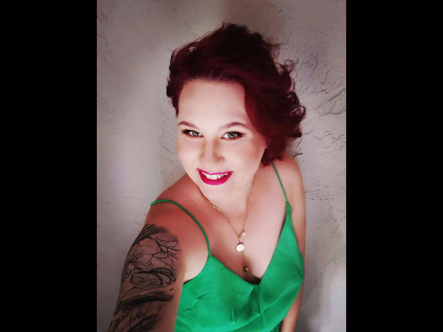 Suniabdsm-hot, com cabelo ruivo encaracolado, vestido verde, braço tatuado e batom rosa, sorrindo para a câmera. Seios visíveis através do vestido. Colar com um pequeno pingente circular..
