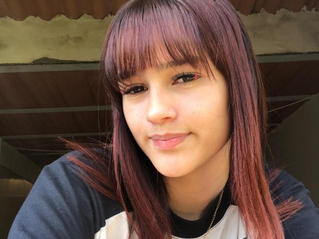 MelodyMansson has straight, red-brown hair with bangs, light skin, and a small smile. She's wearing a black and white shirt with a gold necklace.
