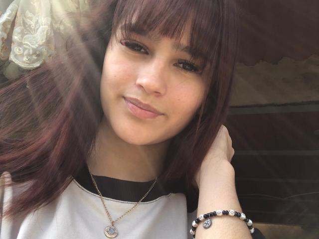 MelodyMansson has brown eyes, light skin, and purple-dyed hair with bangs. She's smiling with a slightly pouty lip, wearing a white shirt and a bead bracelet.