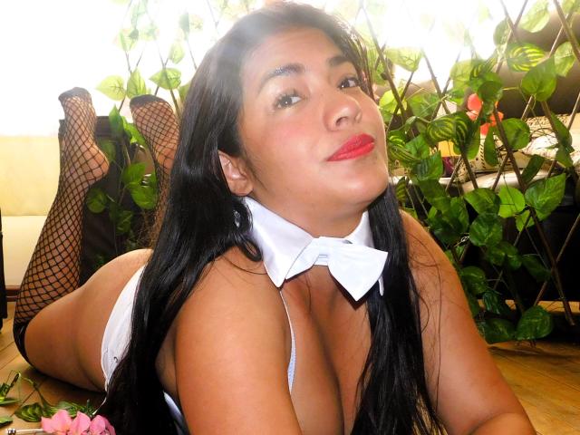 ArianithaXox with long black hair, red lipstick, white bowtie, fishnet stockings, lying on her stomach, showing her tits, ass, and legs, in a sunlit room with plants.