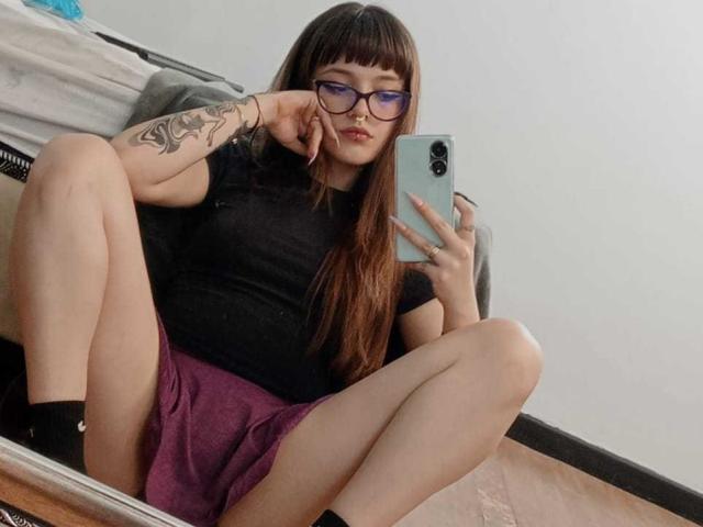 AnniieStone with glasses, black t-shirt, purple skirt, tattooed arm, holding phone, squatting, showing her pussy, long brown hair, black socks, in mirror selfie.
