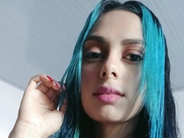 AnaisHilton with teal and black hair, pink lipstick, and red nail polish, looking at the camera with a slight smile. Her hand is near her face.