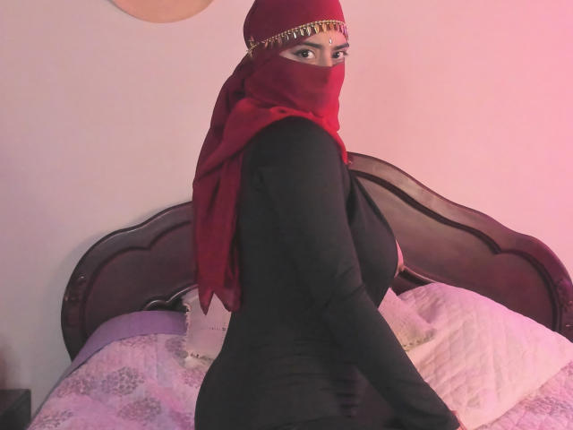 JalinajarN with red headscarf, black dress, and gold chain, looking over shoulder, showing curvy ass, large tits, and a sexy, mysterious expression in a dimly lit bedroom.