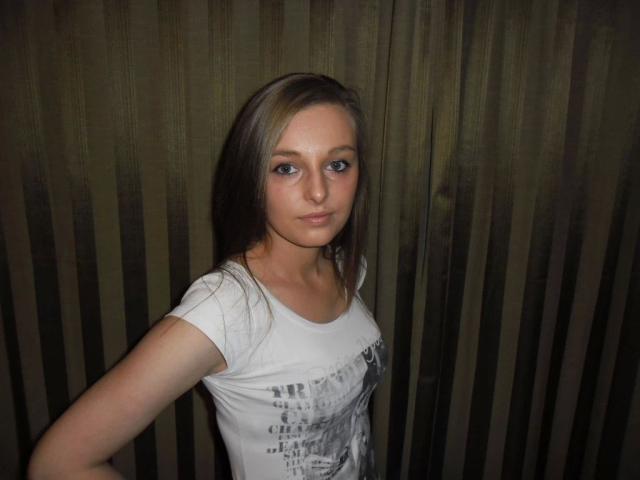 SweetLaila69-hot with long brown hair, blue eyes, white t-shirt, small tits, standing against dark green striped curtain, looking slightly bored, right arm raised.