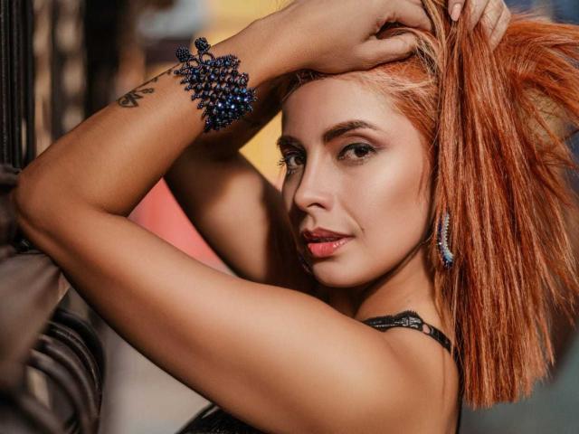 ScarlettElectra mit orangefarbenem Haar, Hände im Haar, trägt ein blaues Perlenarmband, schwarze Ohrringe und einen schwarzen Träger, lächelt in die Kamera, hat ein Tattoo am rechten Arm, helle Haut, sexy..