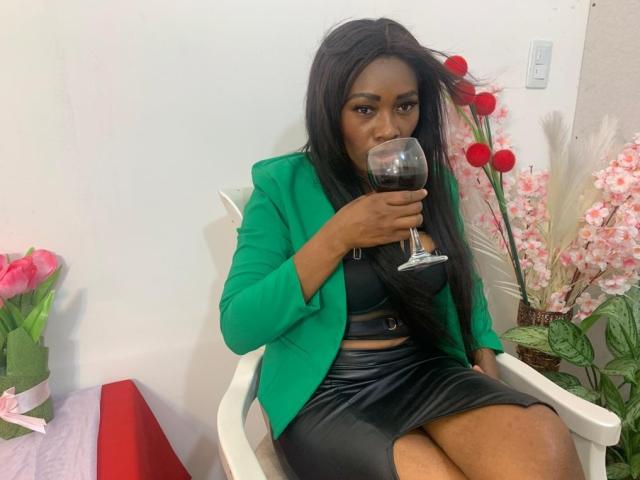AstridDaviz sips red wine, wearing a green blazer over a black bra and shiny black skirt, with long black hair, sitting in a white chair.